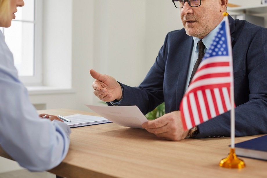usa consul sitting at desk with american flag and talking to woman about visa application